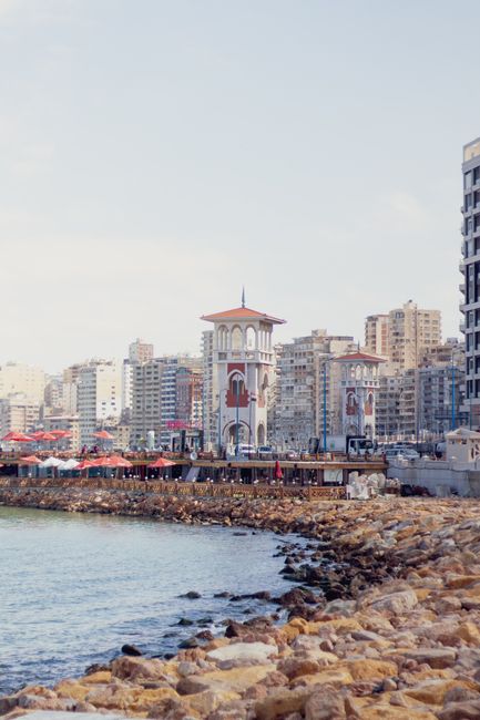 View of Alexandria's Corniche featuring a historic tower and beachfront with city skyline.
