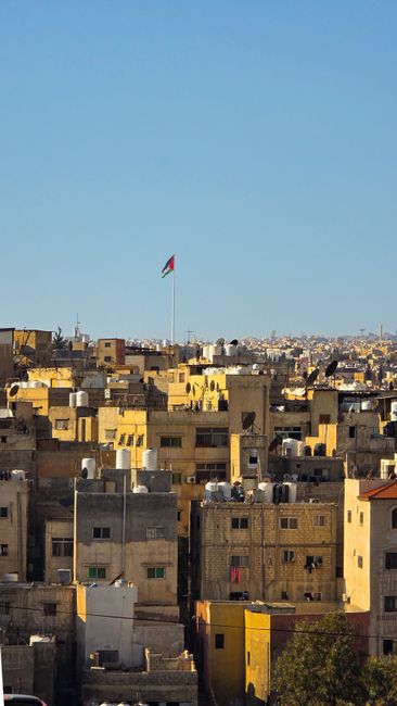 Aerial view of Amman's urban landscape with the Jordanian flag prominently displayed on a sunny day.