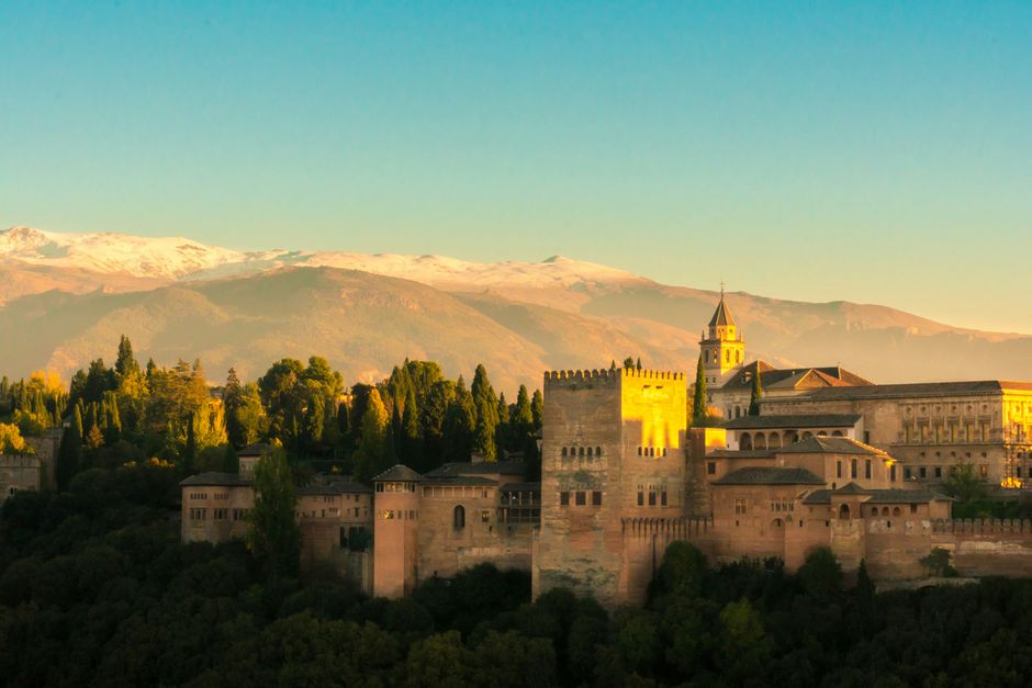 View of Alhambra Palace surrounded by trees with Sierra Nevada mountains in the background at sunset.