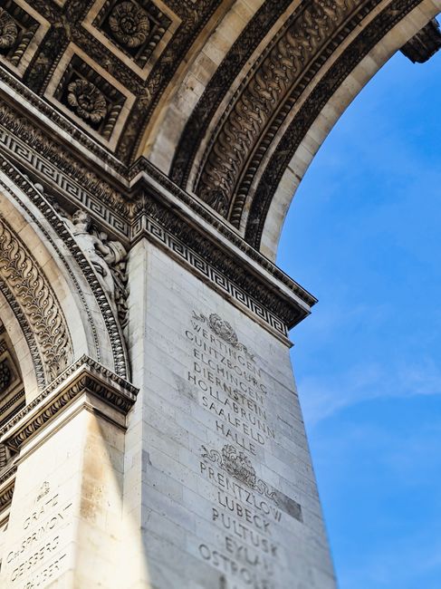 A low-angle view of the Arc de Triomphe in Paris, showcasing detailed carvings against a clear blue sky.