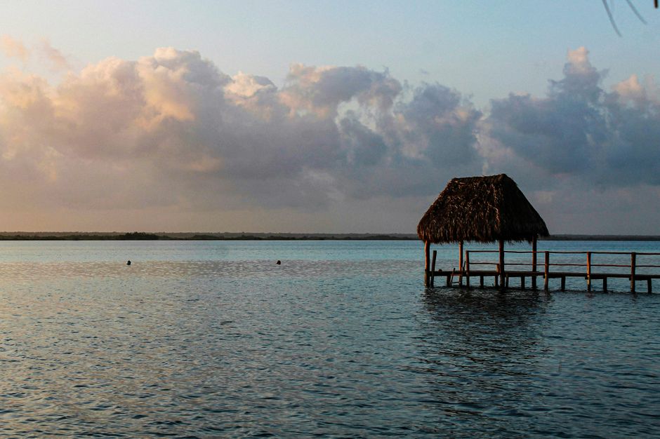 A peaceful view of the Bacalar lagoon at sunrise, featuring a rustic pier and vibrant clouds.