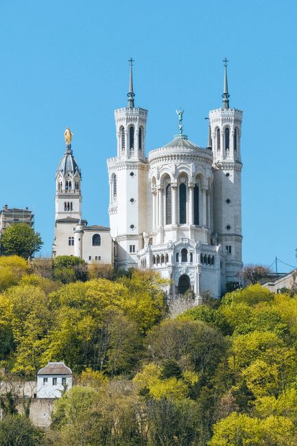 Aged Basilica of Notre Dame de Fourviere exterior surrounded by lush green trees on hill under blue sky in Lyon