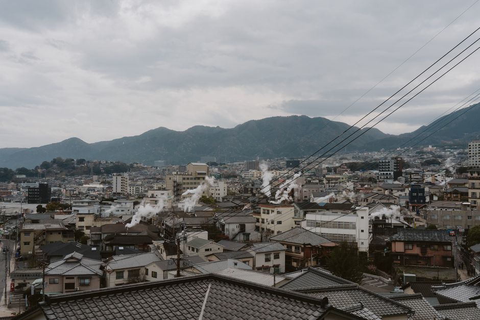 Cityscape view with smoking rooftops and mountain backdrop under overcast skies.