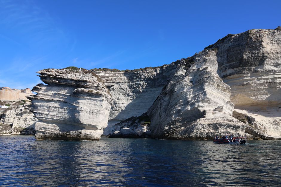 Rugged limestone cliffs of Bonifacio, Corsica, with tourists boating beneath a clear blue sky.