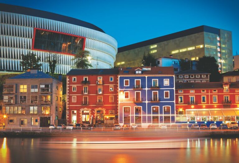 Colorful riverside buildings and San Mamés Stadium in Bilbao illuminated against the night sky.