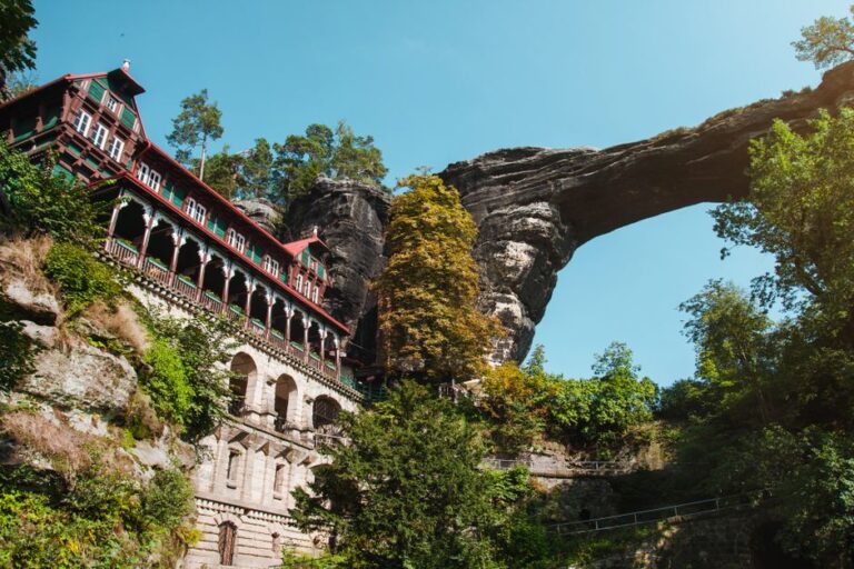 Breathtaking shot of Pravčická Brána, a stunning rock arch, beside a historic building surrounded by lush nature.