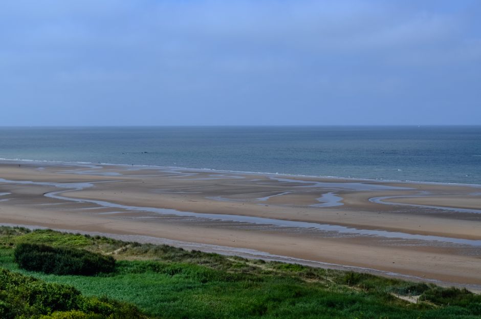 Tranquil view of Omaha Beach in France, showcasing vast sand and calm sea under blue skies.
