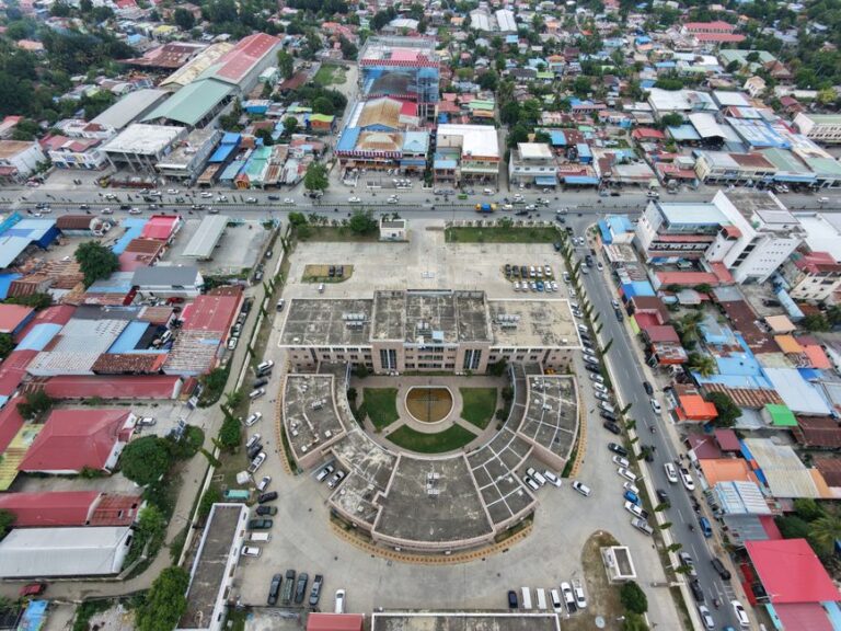 Aerial shot of a cityscape featuring a central building with circular architecture, surrounded by colorful rooftops.