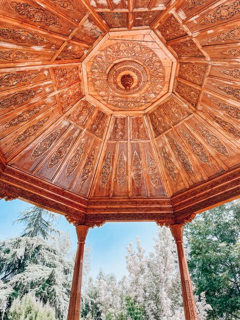 Intricate wooden gazebo ceiling with columns in a lush park setting, Dushanbe, Tajikistan.