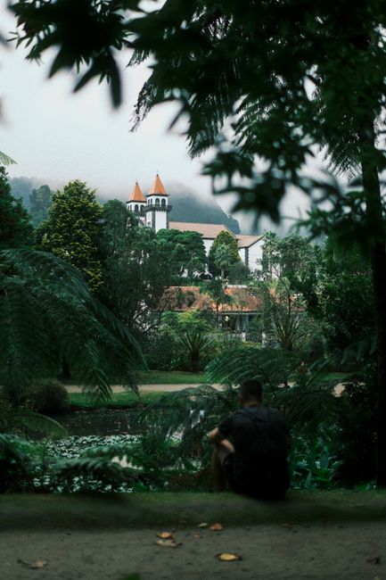 A tranquil scene of a person sitting in Furnas, Azores, Portugal, framed by lush greenery and a historic church in the background.