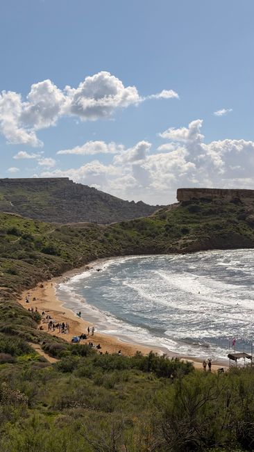 A beautiful view of Golden Bay Beach with waves, hills, and people enjoying the sun in Malta.
