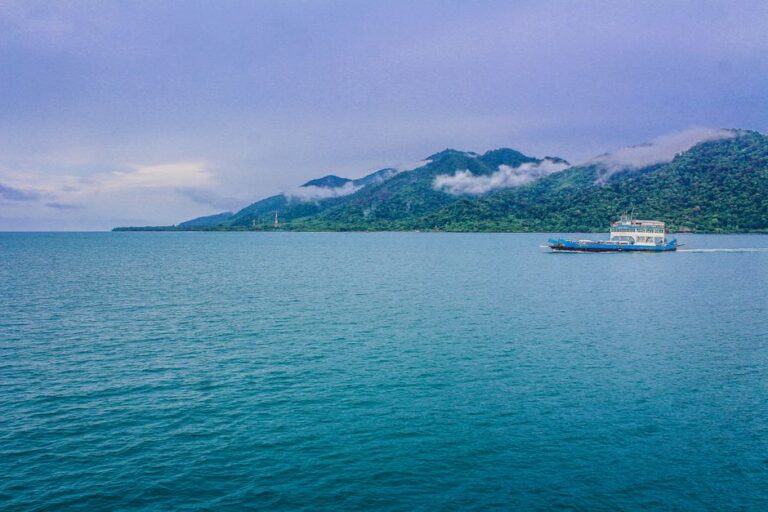 A ferry travels across a serene sea with lush green mountains and clouds in the background.
