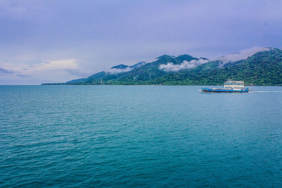 A ferry travels across a serene sea with lush green mountains and clouds in the background.