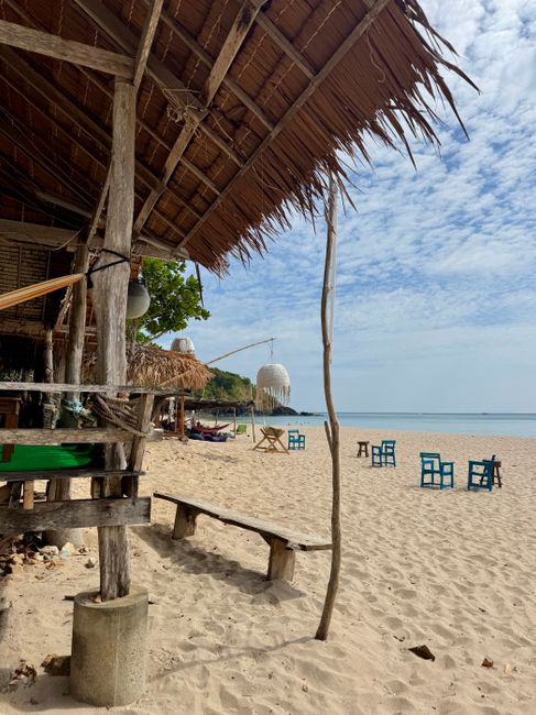 A rustic beach shelter with chairs on a sandy shore under a cloudy sky, evoking a serene seaside atmosphere.