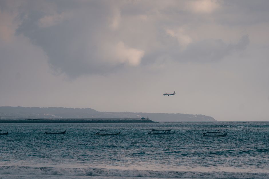 Airplane descends over the scenic coastline of Kuta, Bali, with traditional boats on the sea.