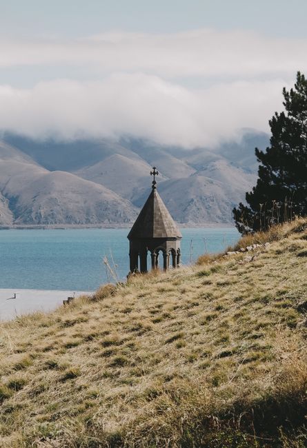 Explore a tranquil view of an ancient church tower near a beautiful lake surrounded by mountains.