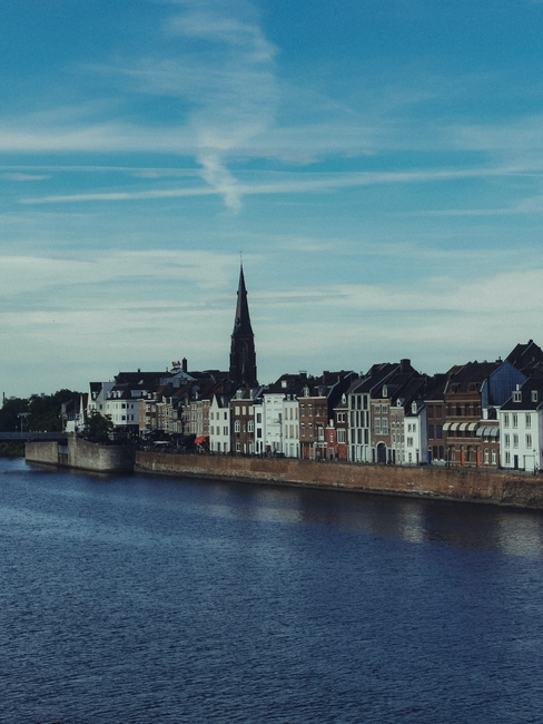 Scenic view of historic buildings along a river with a church spire under a blue sky.