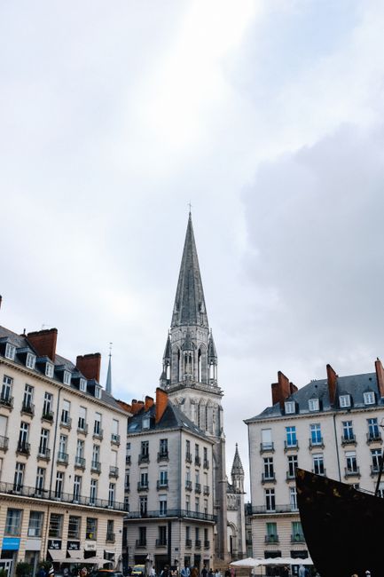 Gothic cathedral tower framed by classic buildings under a cloudy sky in a city setting.