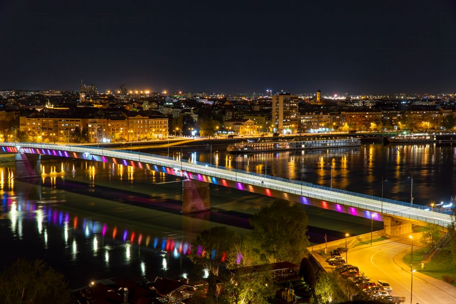 Night view of the illuminated bridge over the Danube River in Novi Sad, showing vibrant reflections and cityscape.