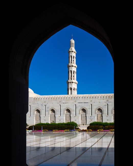 A beautiful view of the Sultan Qaboos Grand Mosque framed by an arch against a clear blue sky.