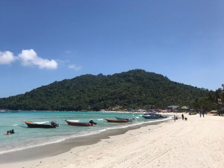 Long-tail boats on a sunny beach with lush hills on the Perhentian Islands in Malaysia.