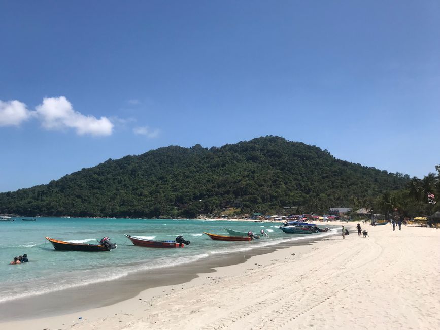 Long-tail boats on a sunny beach with lush hills on the Perhentian Islands in Malaysia.