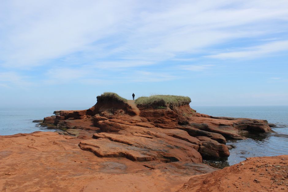 A lone figure stands on red rock cliffs overlooking a calm sea.
