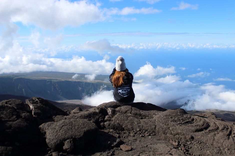A woman sits on a rocky cliff, gazing at the clouds and ocean on Réunion Island.