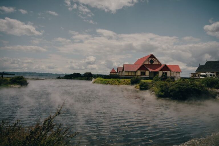 Traditional Maori building by Rotorua Lake with geothermal steam rising, captured in daylight.