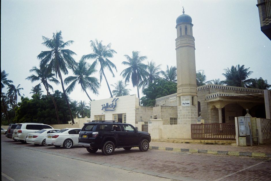 Exterior view of a mosque in Salalah, Oman, with palm trees and parked cars in the background.