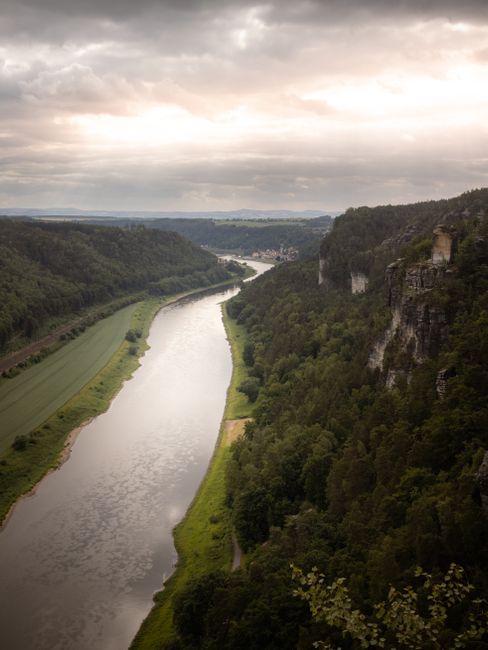 A stunning landscape of the Elbe River winding through the Saxon Switzerland National Park under a dramatic sky.