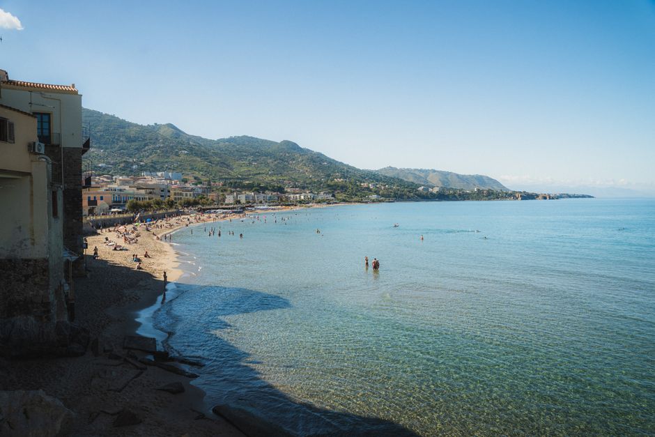 Idyllic coastal scene with a beach, mountains, and clear blue sea under a sunny sky.