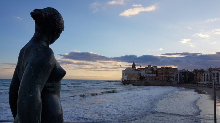 A serene view of the Sitges coastline with a statue in the foreground during sunset.