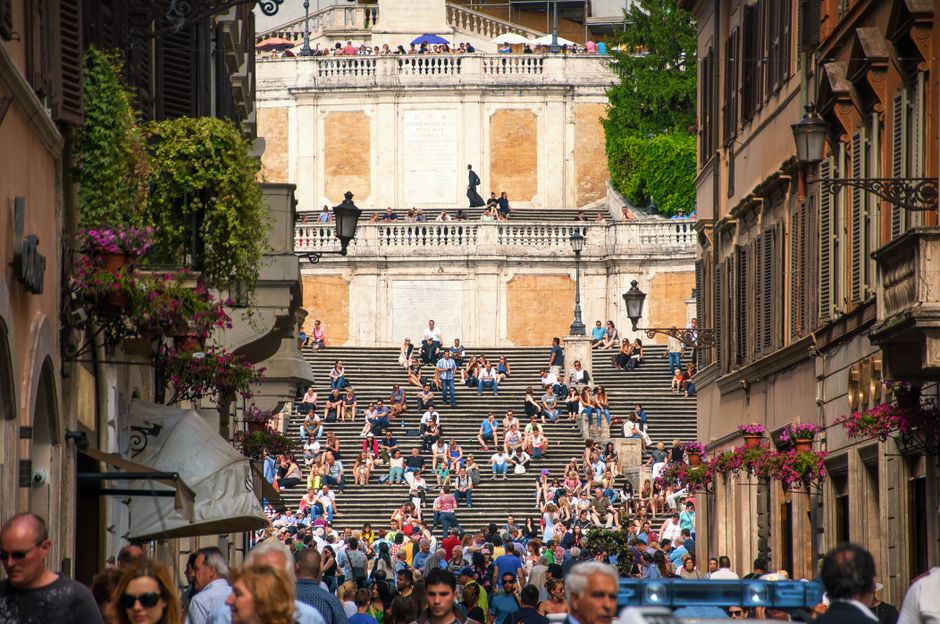 Group of tourists strolling on narrow street and resting on stairs near aged buildings in summer day in city center