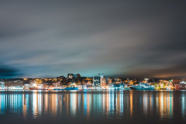 Picturesque scenery of ships floating on sea reflecting glowing lights of St John s city against cloudy evening sky