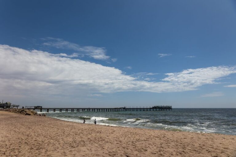 A stunning coastal scene capturing Swakopmund Beach, Namibia, with a pier extending into the ocean under a bright blue sky.