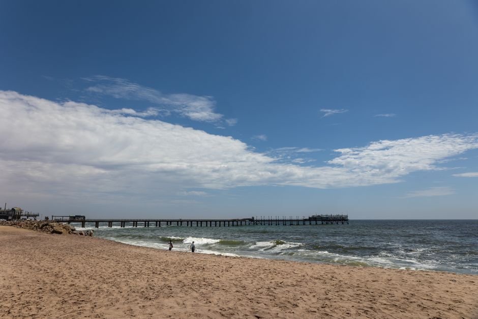 A stunning coastal scene capturing Swakopmund Beach, Namibia, with a pier extending into the ocean under a bright blue sky.