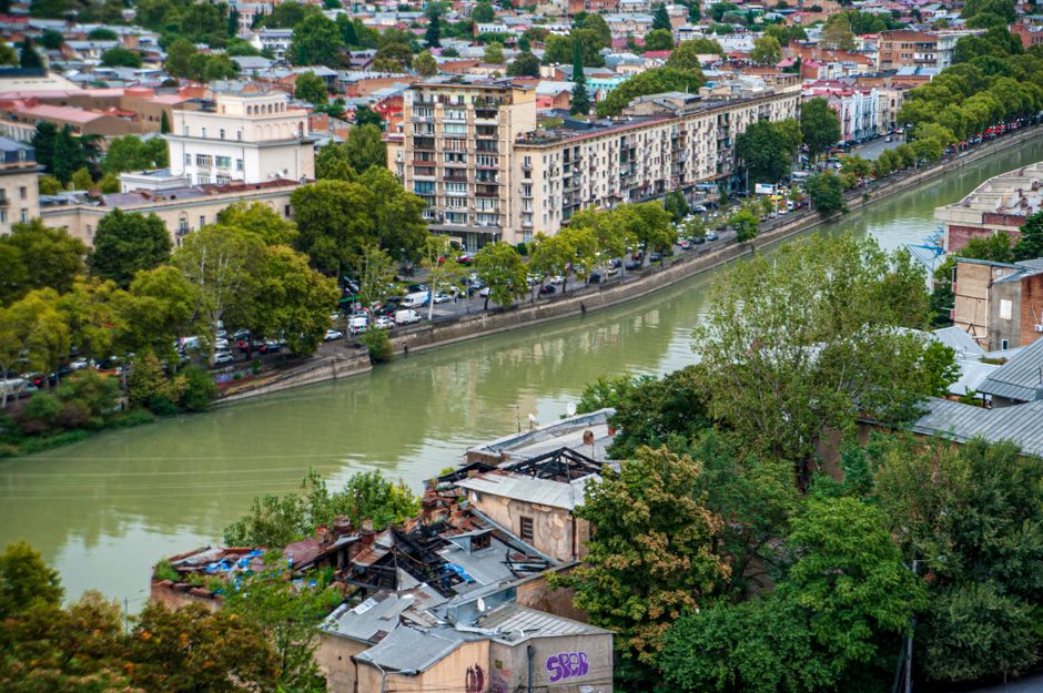 Aerial view of city buildings lining a river surrounded by lush trees and urban scenery.