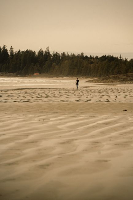 A lone person walks along a serene beach in Tofino, BC, under a soft, overcast sky.