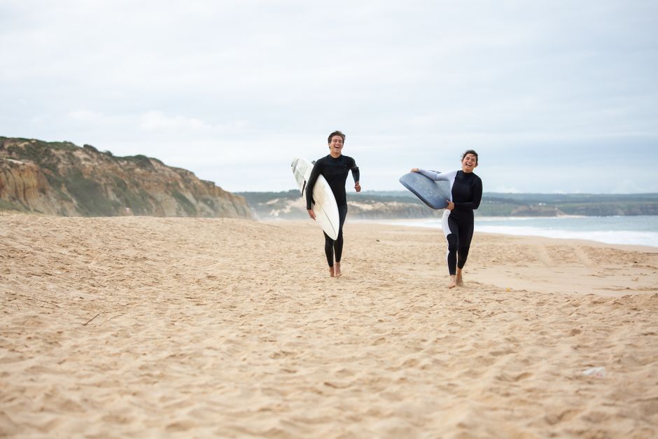 Joyful couple in wetsuits running with surfboards on a picturesque Portuguese beach.