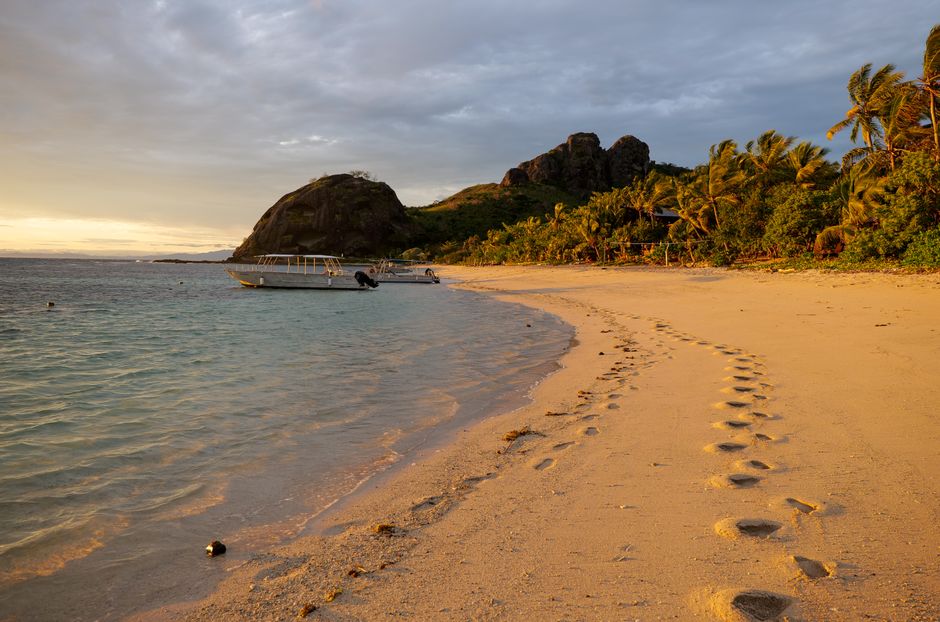 Beautiful beach view of Yasawa Islands, Fiji, at sunset with footprints and boats.